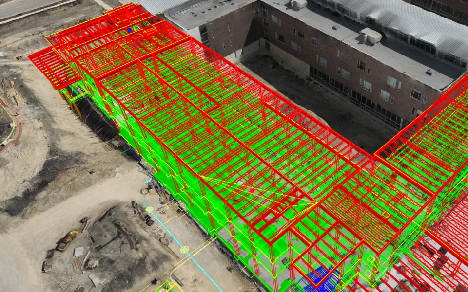 A structural steel model registered onto an aerial capture of the same building under construction, with members colour-coded against the model so erected steel reads green and outstanding steel reads red.