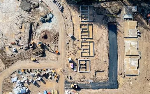Nadir orthomosaic covering a full construction site: stem-wall footings in formwork, poured slab pads, an open utility trench, a haul road and a laydown yard, all at one constant scale.